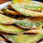 A close-up of crispy avocado chips dusted with seasoning, stacked in a white bowl, offering a crunchy, golden texture.