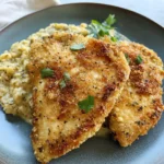 Two crispy baked chicken cutlets garnished with parsley, served over a creamy risotto-style dish on a dark plate.
