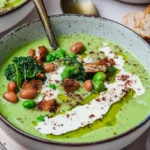 A bowl of green soup with white beans, peas, kale, and crispy bread pieces, drizzled with cream and olive oil.